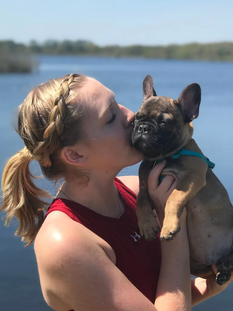 A woman kissing Charlie as a puppy, in his teal harness beside a lake